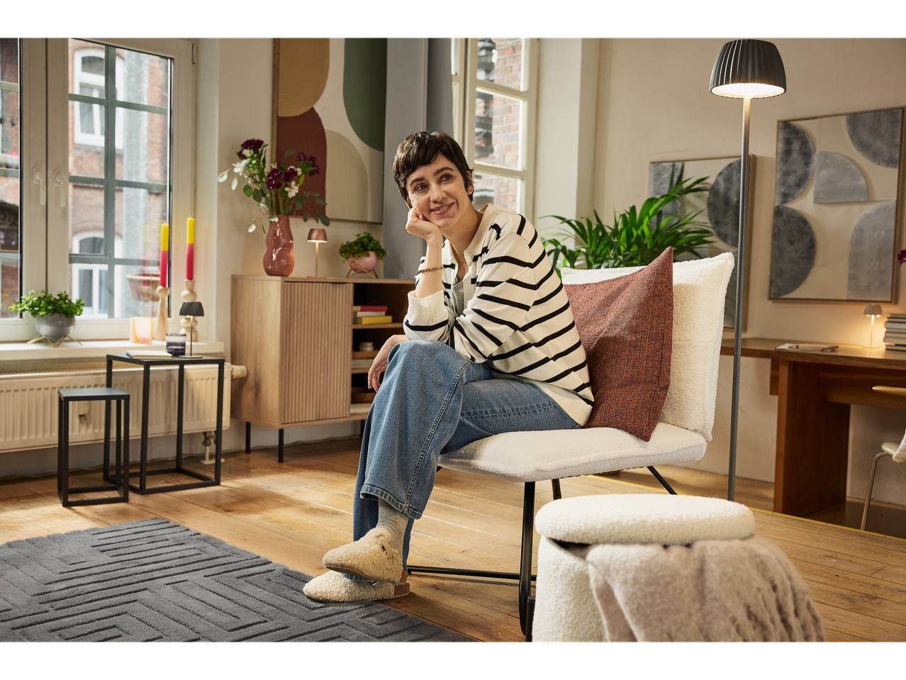 Mulher sentada em cadeira com arrumação, rodeada por mobiliário e decoração modernos.