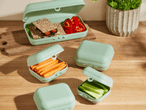 Hellgrüne Brotdosen mit Sandwiches, Paprika, Karotten und Gurken auf einem Holztisch.