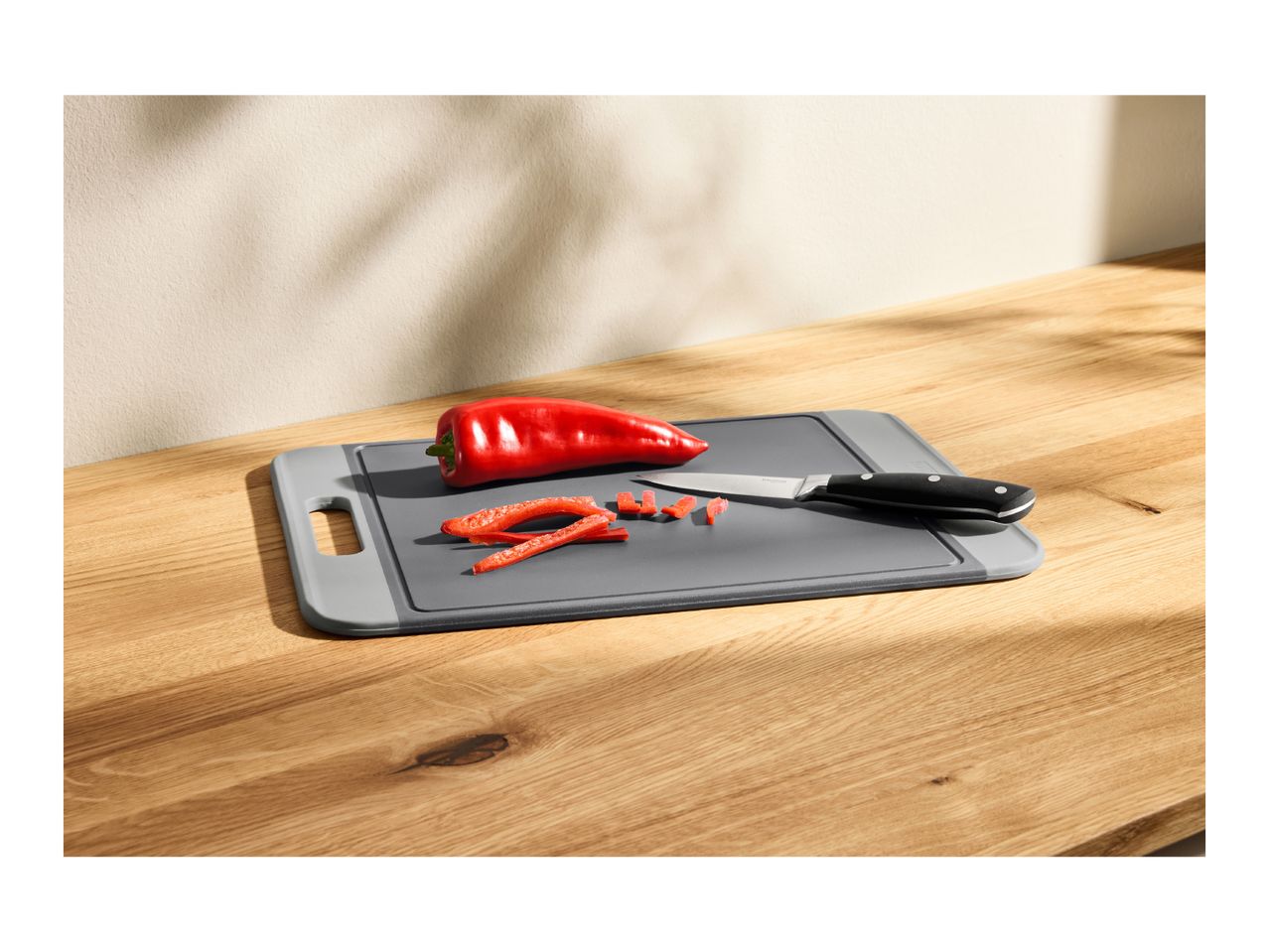A grey plastic cutting board with a red pepper and a knife on a wooden table.