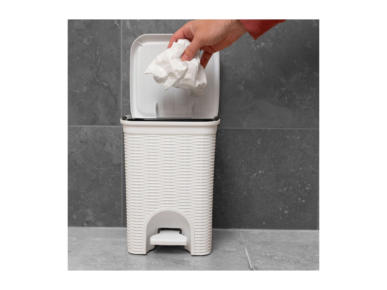 Hand dropping crumpled paper into a white pedal bin with a woven texture, against a grey tiled background.