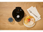 A SilverCrest waffle maker sits on a wooden table next to a plate of waffles and a bowl of blueberries.