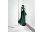 A green folding hand truck is stored upright in a corner against a white wall.