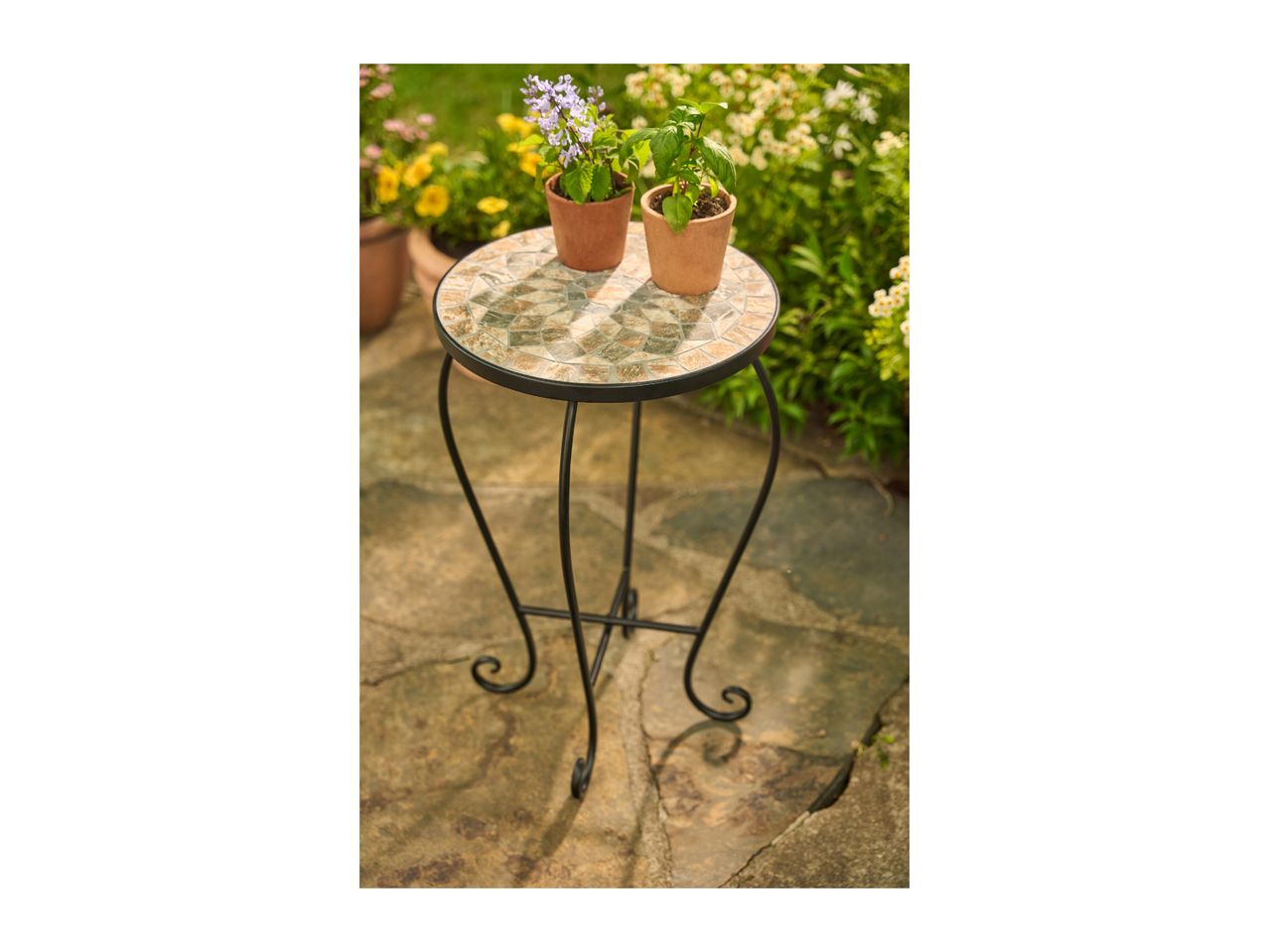 Round mosaic garden table with two potted plants on a patio, surrounded by flowers.
