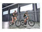 Two people riding electric bikes in an indoor parking garage with bright windows.