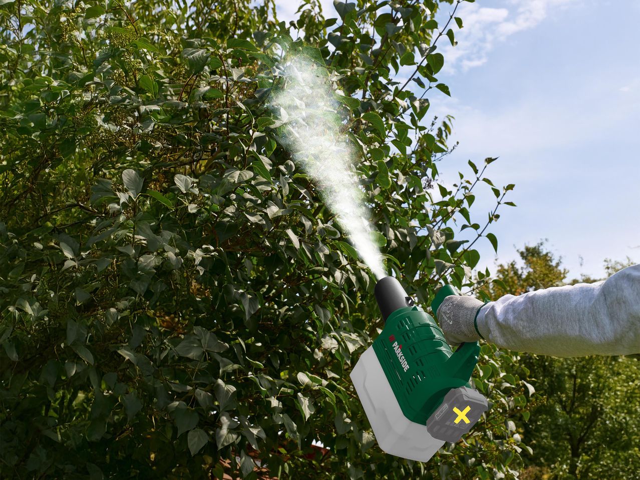 Egy személy permetezőgéppel permetez egy bokrot, a permetezőgépből fehér permet áramlik ki.