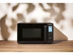 A black microwave oven with a digital display on a wooden table, with shadows on the wall.