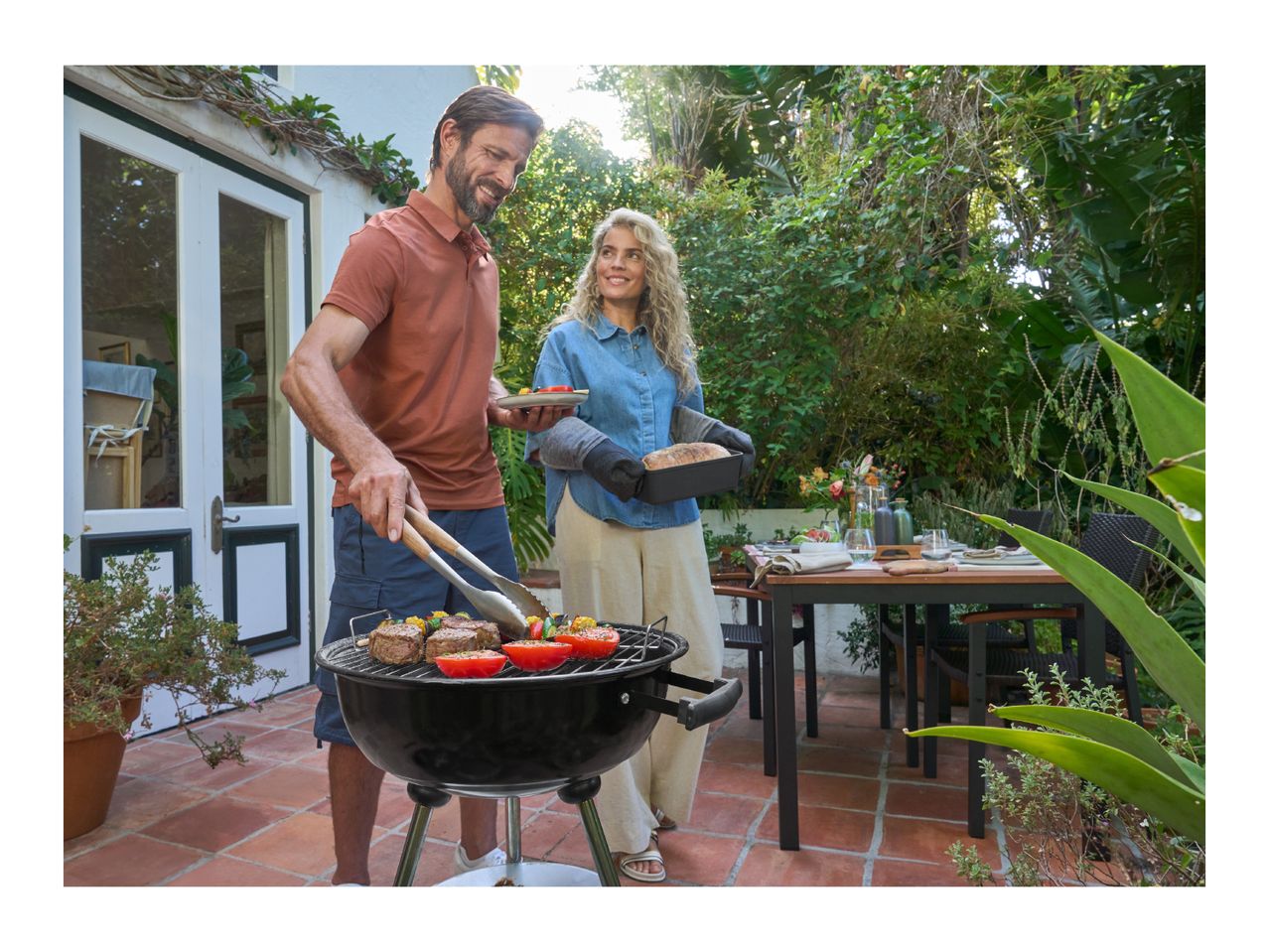 Man grilling meat and vegetables, woman holding a loaf of bread, outdoor dining.