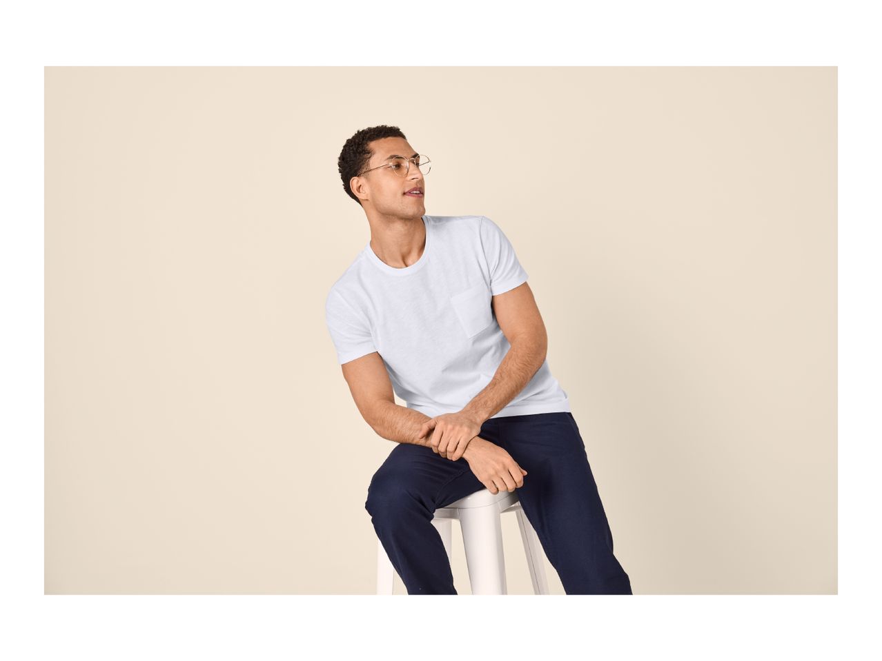 Man in white t-shirt and dark blue trousers sitting on a stool, looking right.