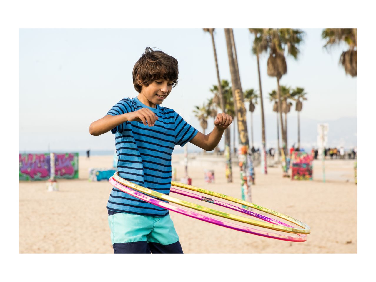 A boy in a blue striped t-shirt and swim shorts hula hooping on a sandy beach.