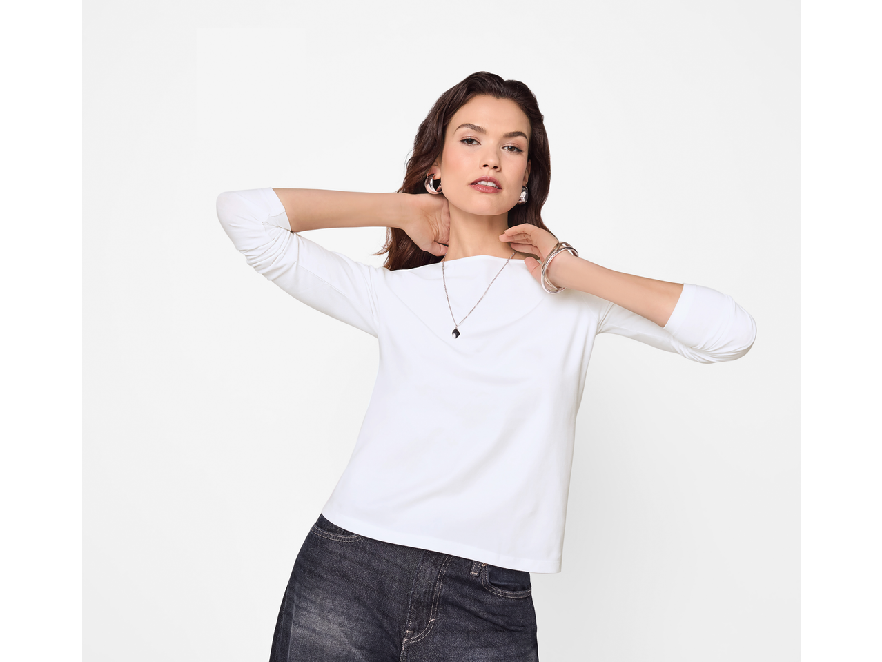 Woman wearing a white esmara® long sleeve top and dark jeans, posing with hands behind her neck.