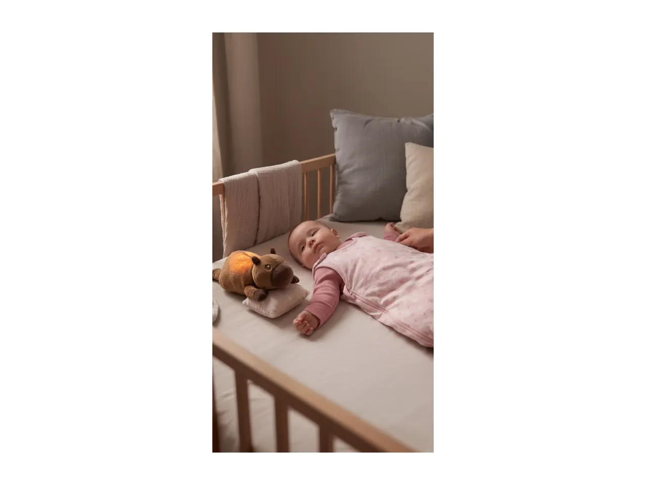 Baby in a pink sleepsuit lying in a wooden cot next to a glowing plush toy.