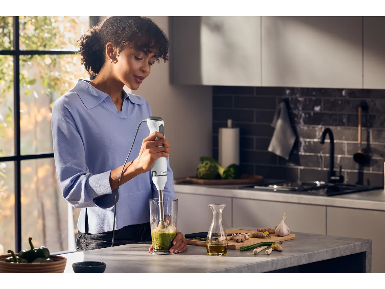 Woman blending green ingredients in a glass with an immersion blender in a modern kitchen.