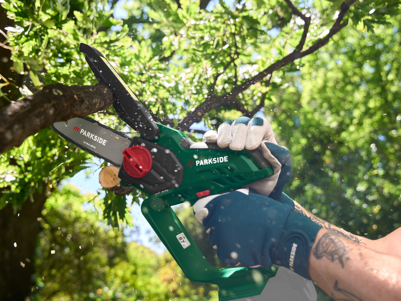 Άτομο με γάντια κλαδεύει ένα κλαδί δέντρου με ένα πράσινο μίνι αλυσοπρίονο, με πριονίδι να πετάγεται.