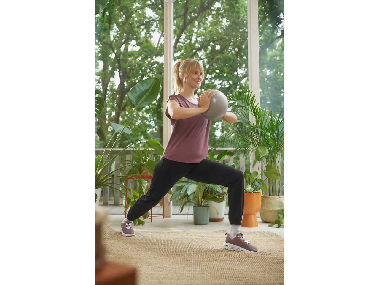 Moteris su Crivit sportbačiais ir marškinėliais, daro įtūpstus su pilates kamuoliu.