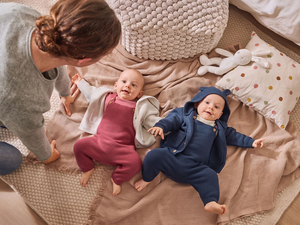 A person interacting with two babies dressed in knit rompers and cardigans on a blanket.