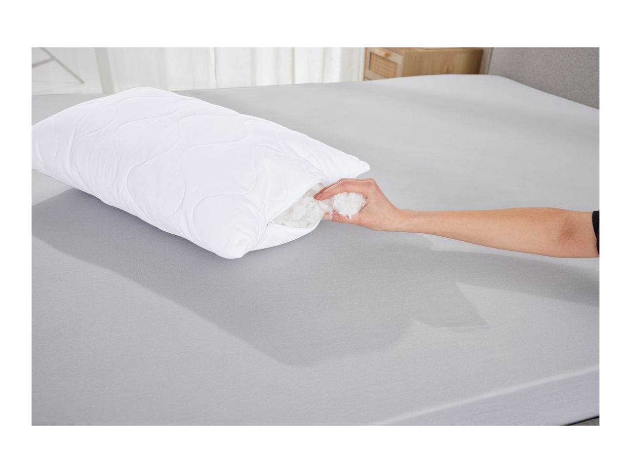A hand adjusting the filling of a white quilted pillow on a grey bed.