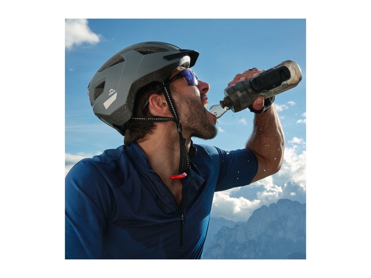 Cyclist in helmet and sunglasses drinking from a water bottle with mountains in background.