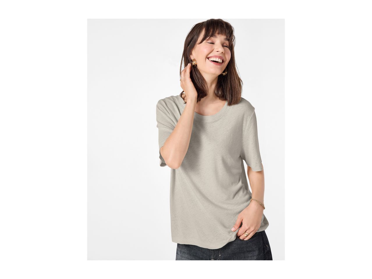 Smiling woman in a light grey t-shirt and dark jeans, with gold earrings and bracelet.