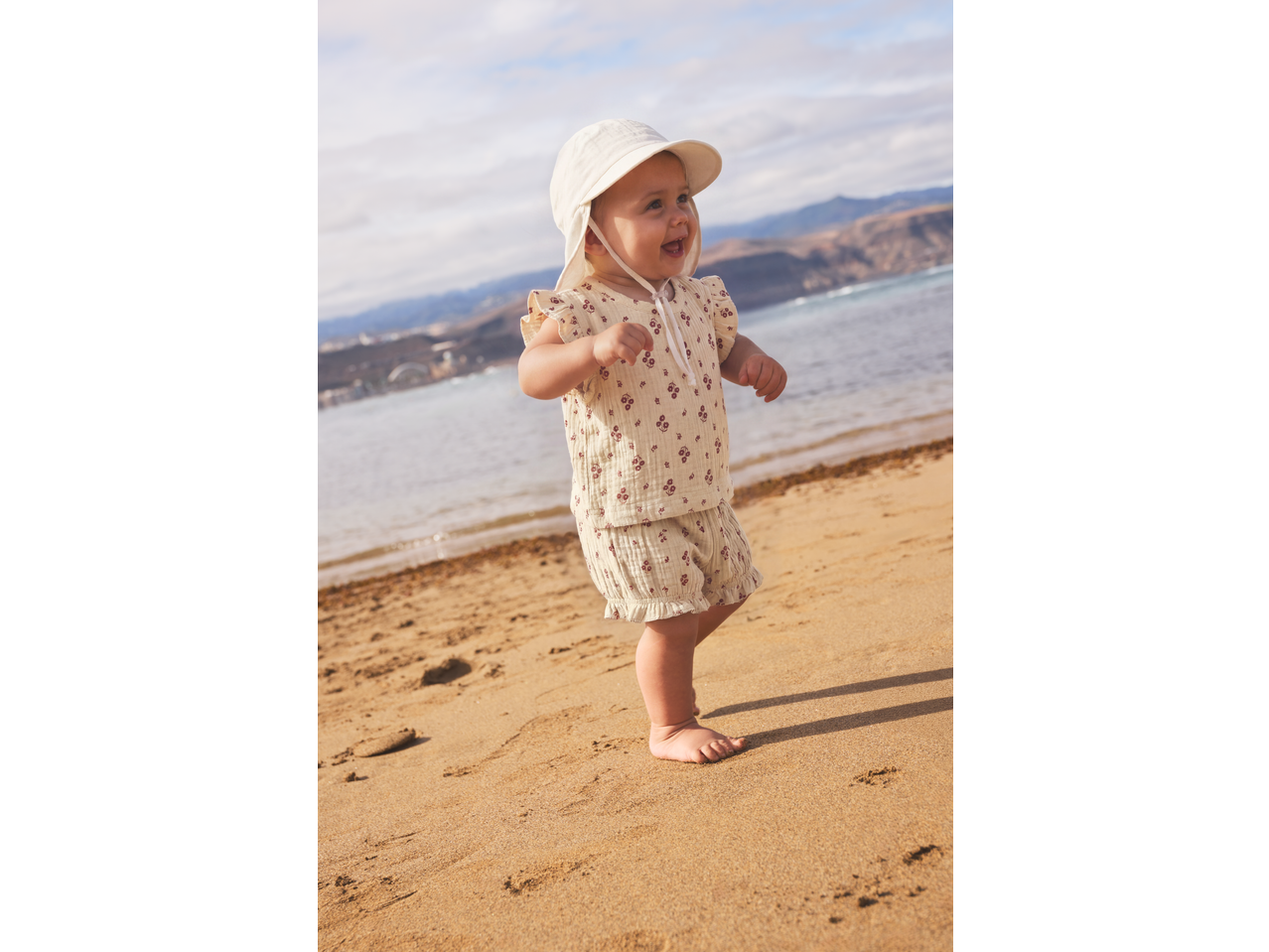 Happy baby in a sun hat and floral outfit walking on a sandy beach.