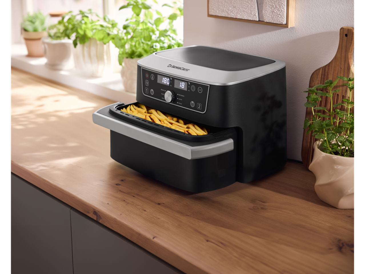 Heißluftfritteuse mit Pommes auf einer Holzarbeitsplatte, umgeben von Topfkräutern.