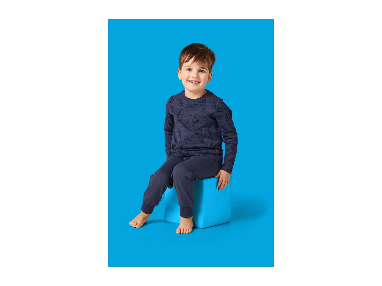 Smiling boy in dark blue pajamas with construction vehicle print, sitting on a blue cube.