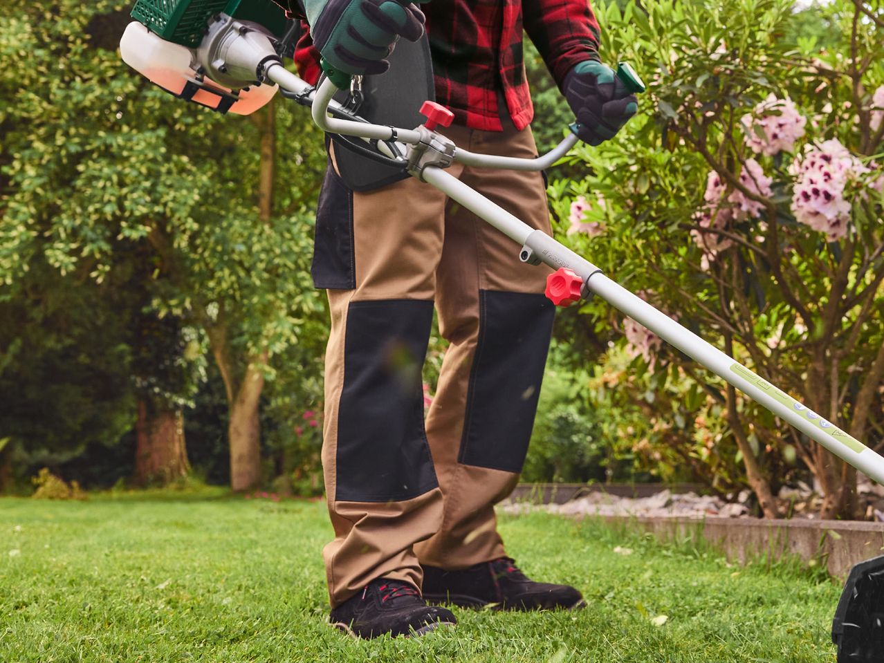 Person in workwear using a gas-powered brush cutter in a garden.