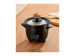 Black rice cooker with cooked rice inside, on a wooden counter next to a cutting board and cloth.