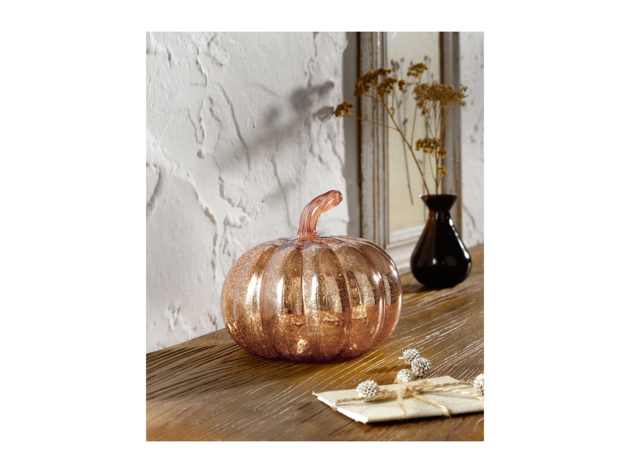Rose gold glass pumpkin on a wooden surface with a dark vase and dried flowers.