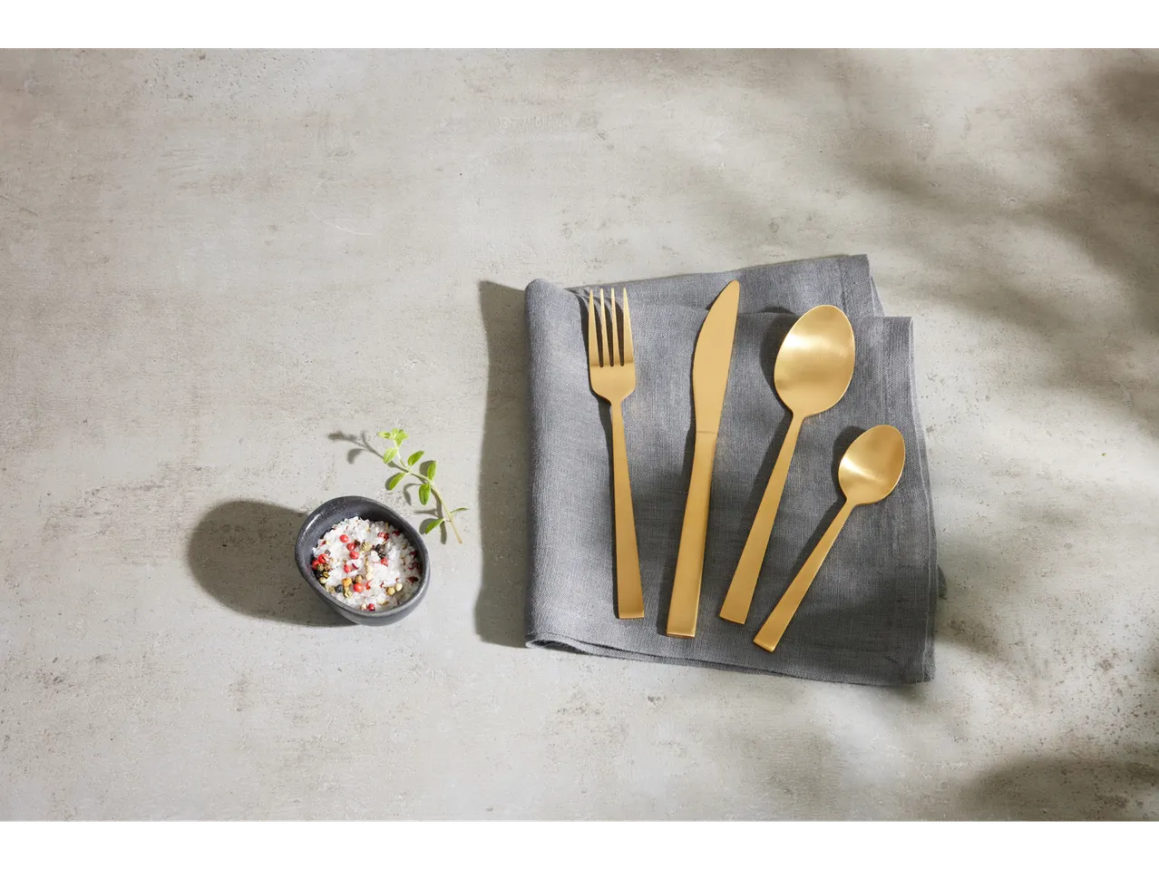 Gold stainless steel cutlery set on a gray linen napkin with a bowl of salt.