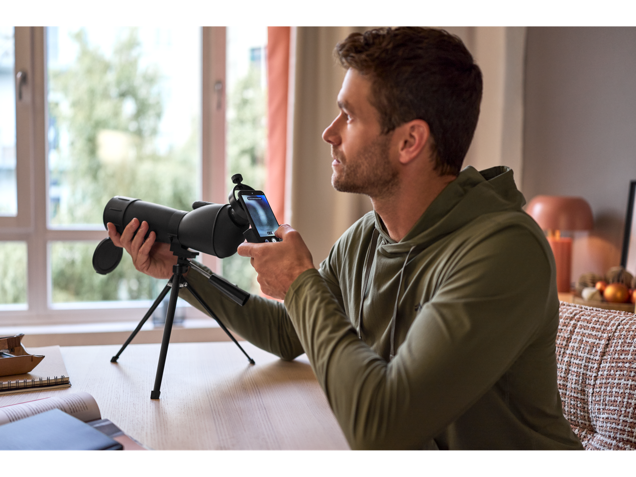 Homem a usar CRIVIT® Spektiv com adaptador para smartphone junto a uma janela