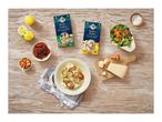Two risotto mixes, one with vegetables and one with almonds, zucchini, orange, and lemon, on a wooden table with ingredients and a prepared dish.