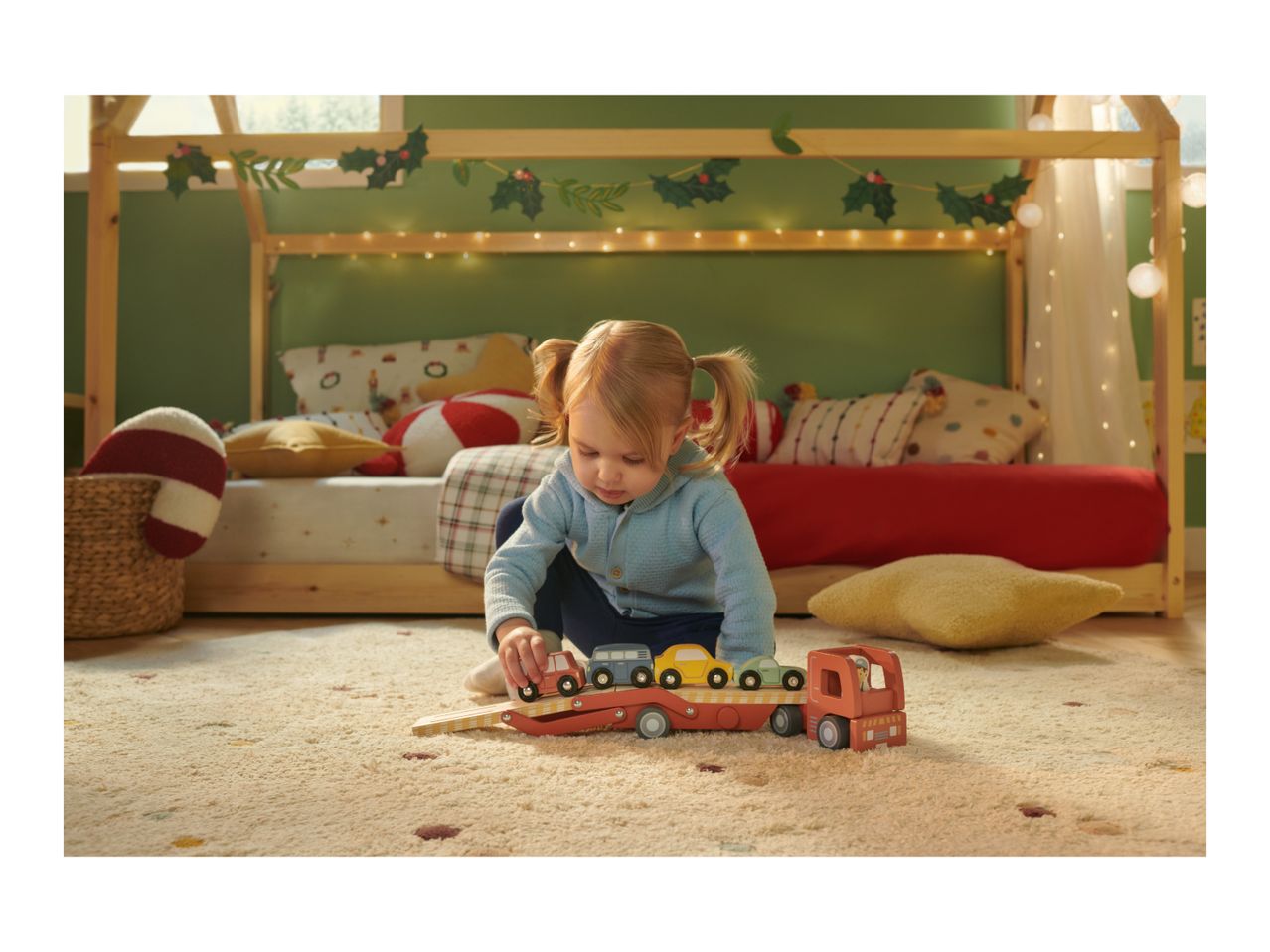Toddler playing with a wooden car transporter toy on a rug in a festive bedroom.