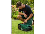Man kneeling on grass, operating a robotic lawnmower in a garden.