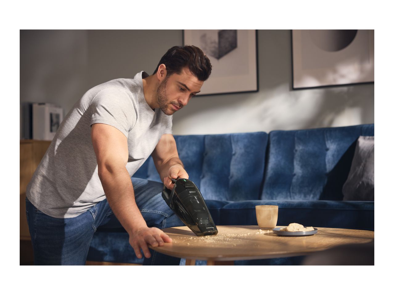 Man cleaning crumbs from a wooden coffee table with a handheld vacuum cleaner.