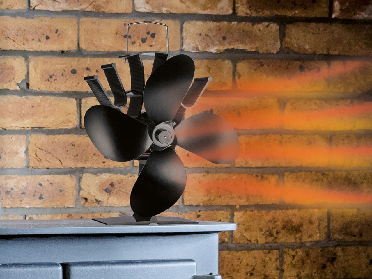 Black heat-powered stove fan on a stove, with warm air spreading across a brick wall.