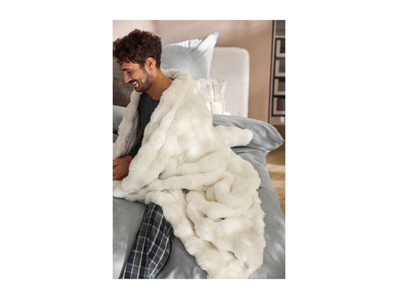 A man sitting on a bed wrapped in a soft, white faux fur blanket.