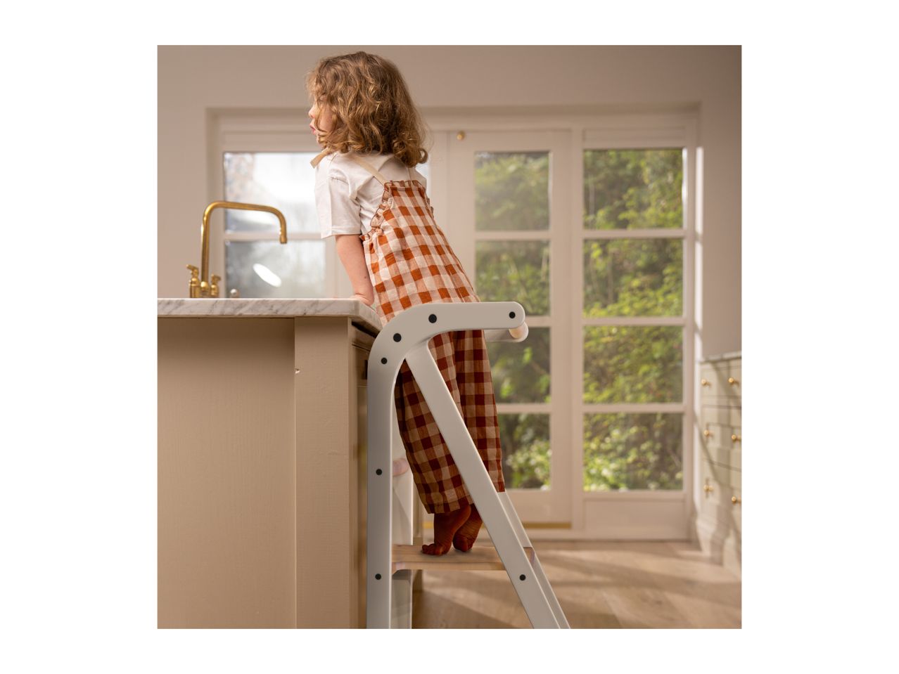 Child on a learning tower in a kitchen, looking at a golden faucet.