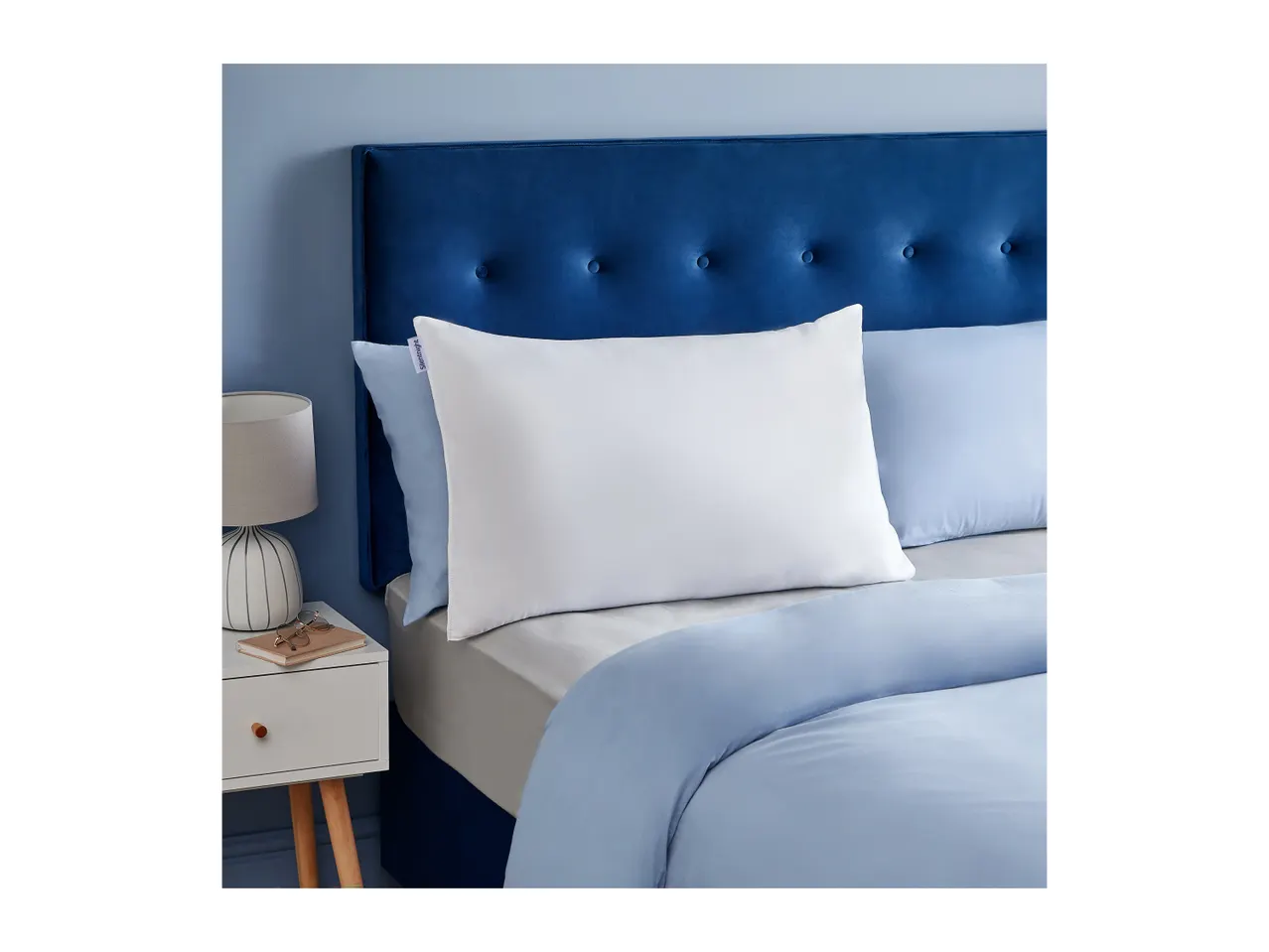 A white pillow with a Silentnight tag on a bed with blue bedding and a blue headboard.