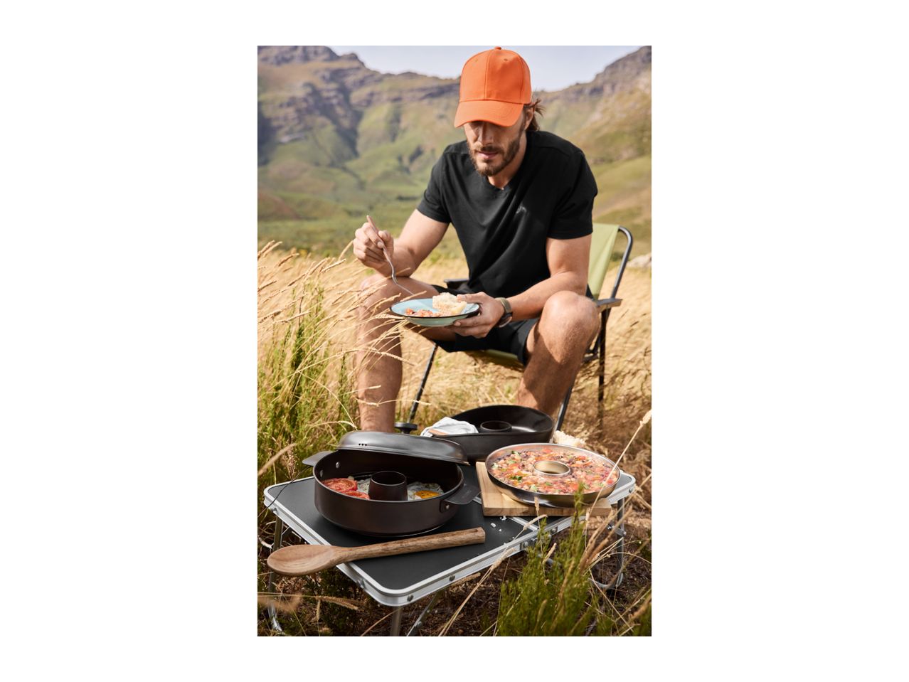 Man eating outdoors from camping cookware on a portable table in a mountainous landscape.