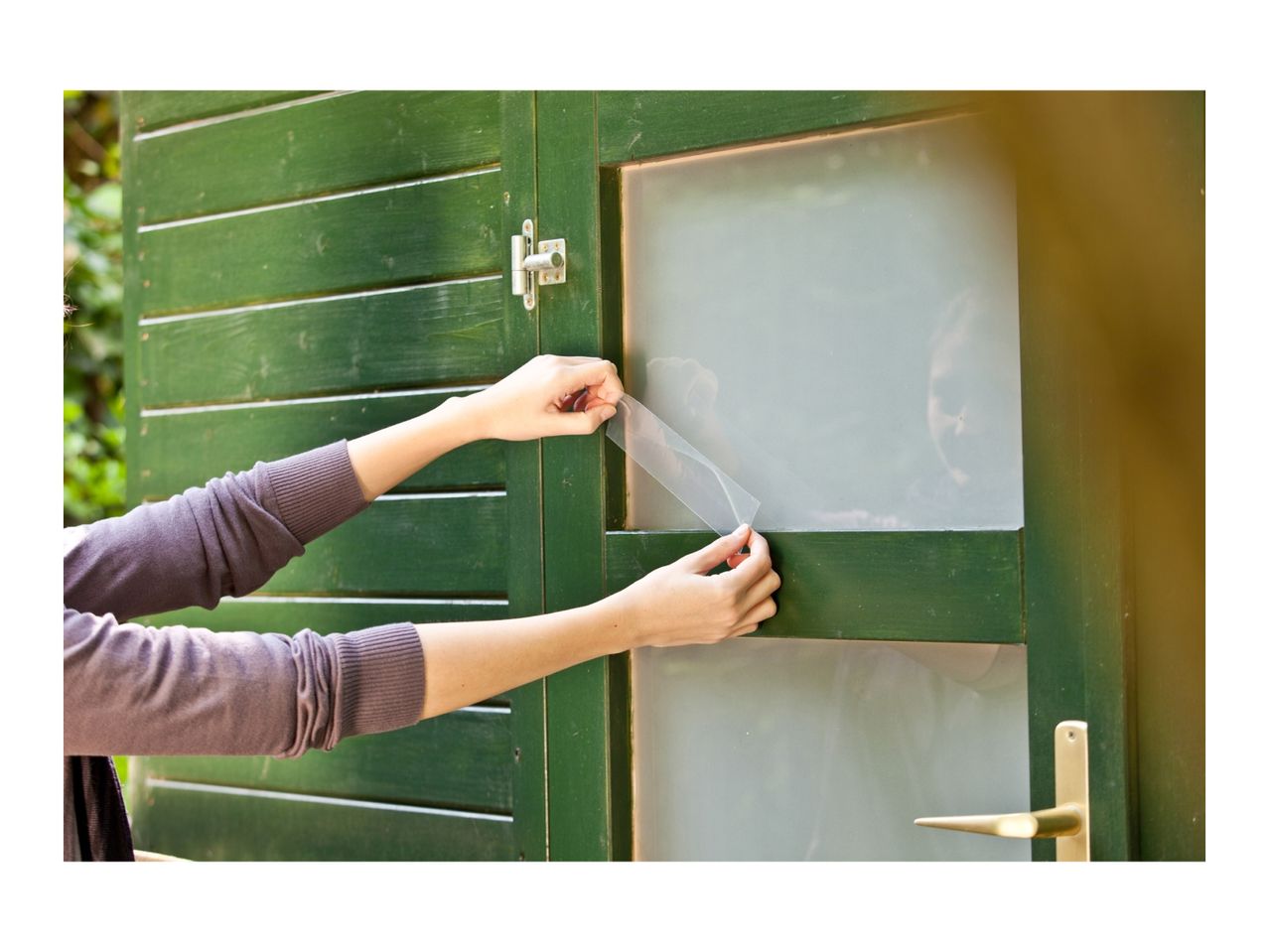 Hands applying privacy film to a window on a green wooden shed.