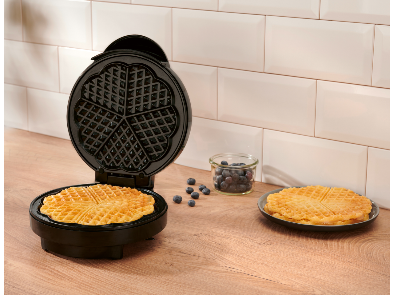 Waffle maker with a fresh waffle inside, blueberries, and a stack of waffles on a plate.