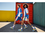 Two models in tracksuits, shorts, socks, and sneakers, walking past colorful shipping containers.