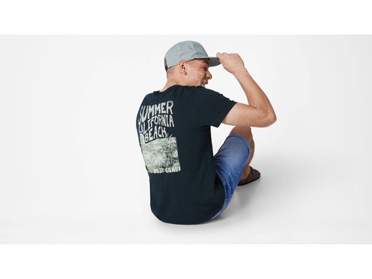 Uomo seduto che indossa una t-shirt nera con stampa e un cappello da baseball.