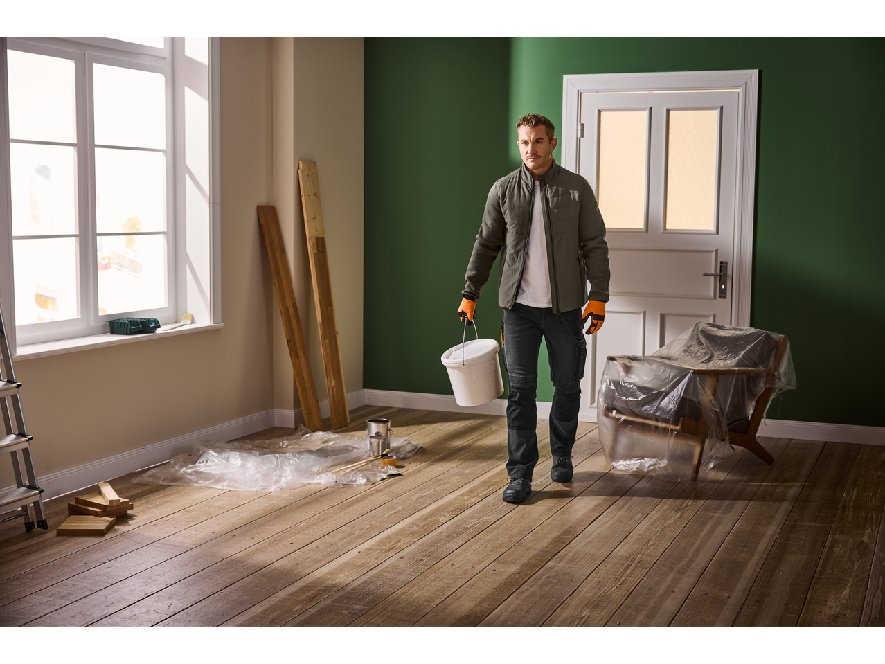 Homem com roupa de trabalho a carregar um balde de tinta numa divisão a ser pintada.