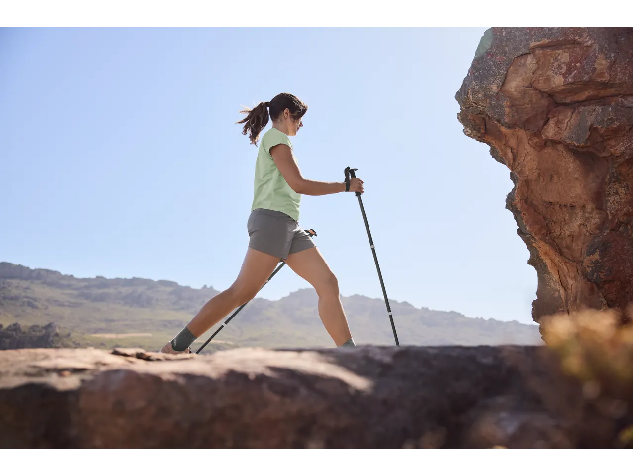Woman in a light green t-shirt and grey shorts hiking with trekking poles.