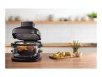 Air fryer with food inside, next to potatoes and rosemary on a wooden board.