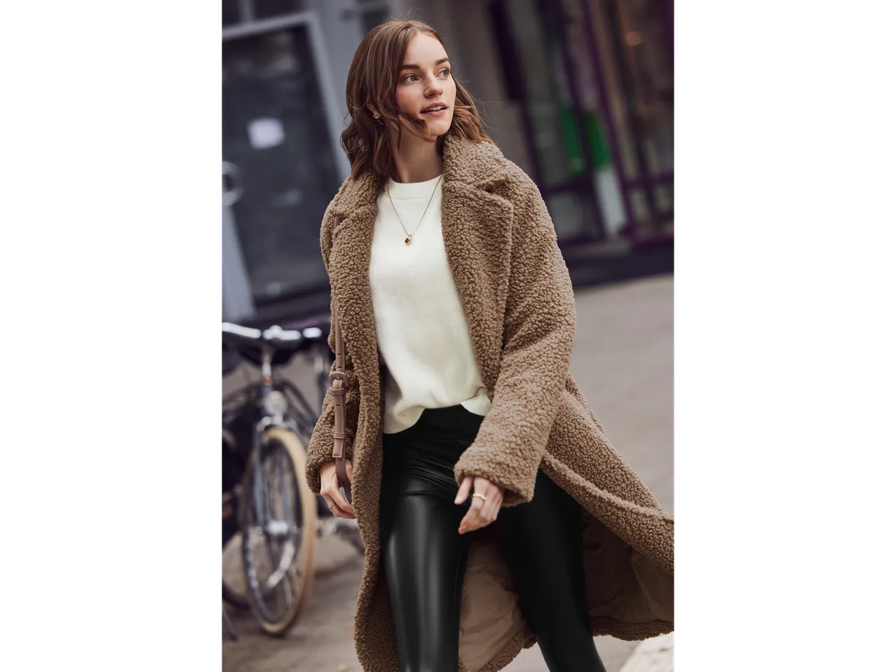 Donna con cappotto teddy marrone, maglione crema e pantaloni in pelle neri.