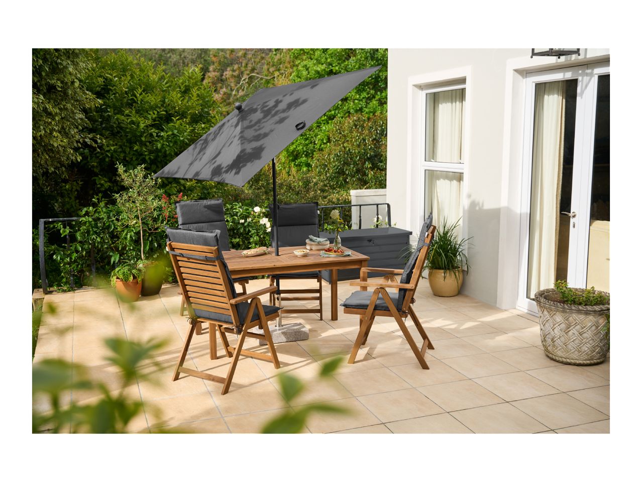 Wooden patio dining set with grey cushions and a large grey parasol on a tiled patio.