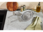 Stainless steel sieves on a kitchen counter with a cutting board and spices.