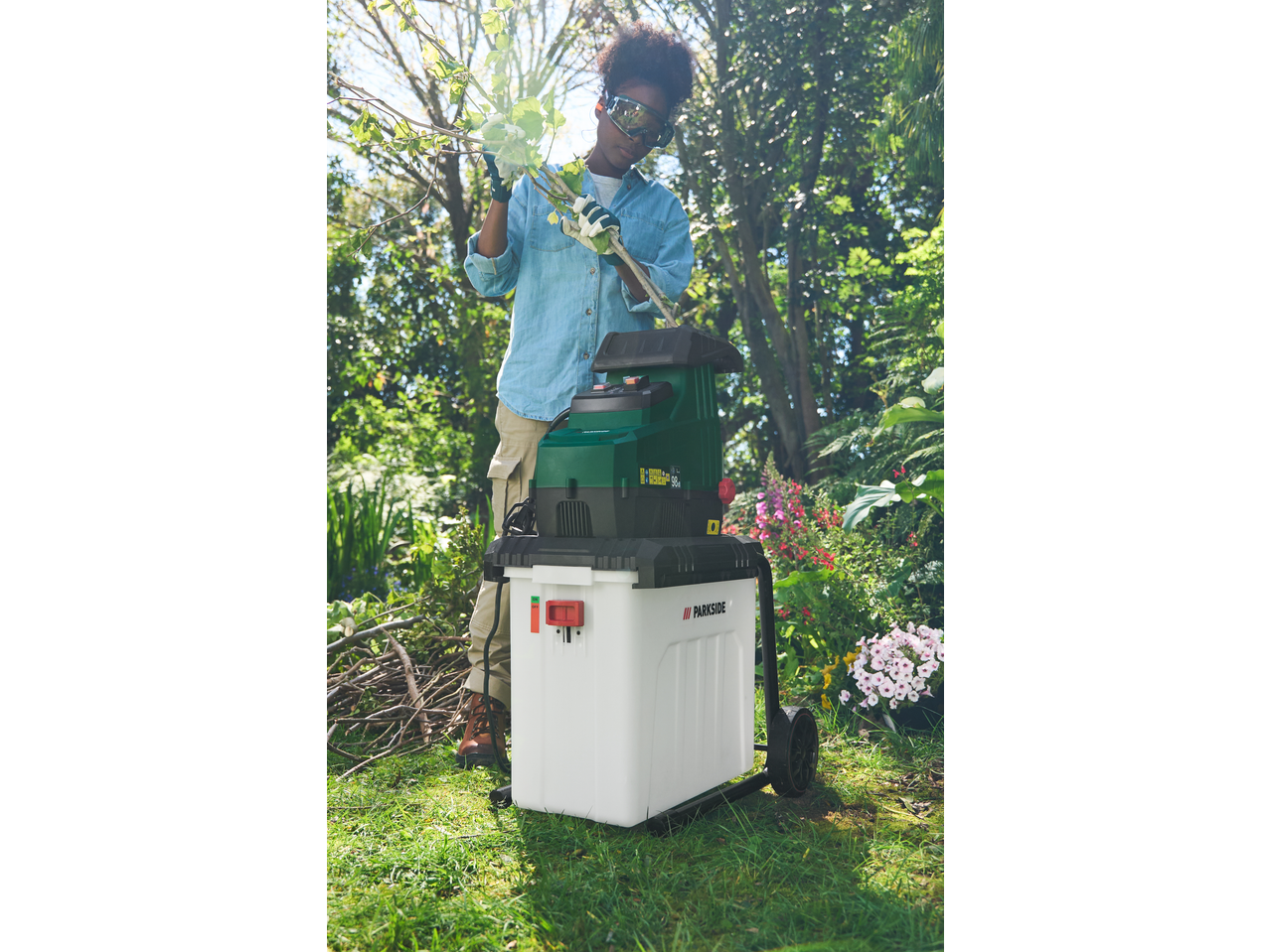 Ženska z zaščitnimi očali in rokavicami uporablja drobilnik PARKSIDE na vrtu.